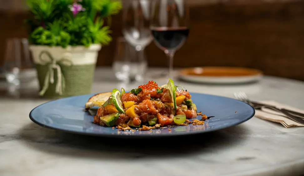 Tartare with mango and avocado on a blue plate, with a glass of red wine behind